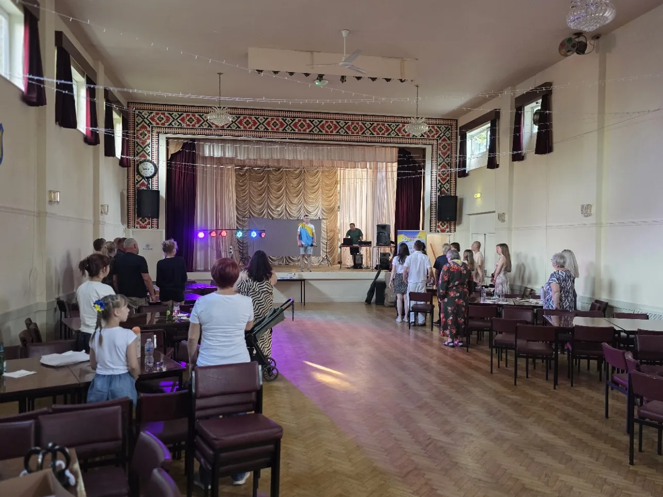 A hall with embroidered pattern decoration around the stage, strings of lights overhead, tables and chairs set out and a performer singing on stage while people stand and watch